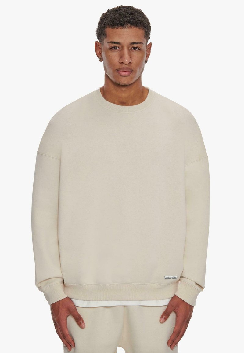 Beige huppari, joka on valmistettu pehmeästä kankaasta. Siinä on pyöreä kaula-aukko, pudotetut olkapäät ja ribbireunukset. Minimalistinen muotoilu, jossa on tarratiedot.