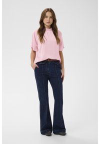 Blusa rosa chiaro a maniche corte con scollo arricciato, abbinata a jeans svasati blu scuro con cuciture verticali e cintura marrone.