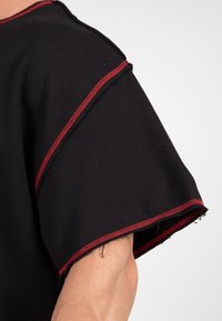 Black garment with wide sleeves, featuring red stitching along seams. Fabric appears textured, with raw edges at the sleeve and hem.