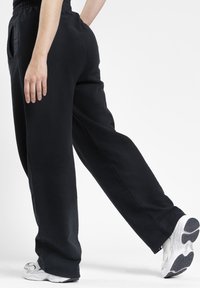 Eine Person, die weite schwarze Hosen und weiße Turnschuhe trägt, geht, von der Taille abwärts vor einem weißen Hintergrund gezeigt.