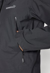 Svart vattentät jacka med en texturerad yta, med fickor med dragkedja, dragkedjehandtag och en logotyp på bröstet.