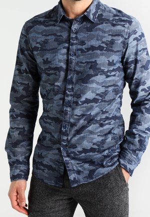 Mann trägt ein blau-graues Tarnmuster-Hemd mit langen Ärmeln und dunkelgraue Hose, Hand in der Tasche.