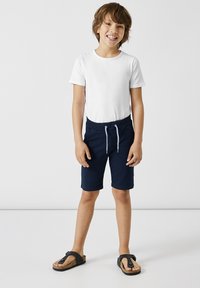 Vit kortärmad T-shirt, marinblå shorts med dragsko, och svarta flip-flops. Enkel design, lättviktig tyg, avslappnad stil.