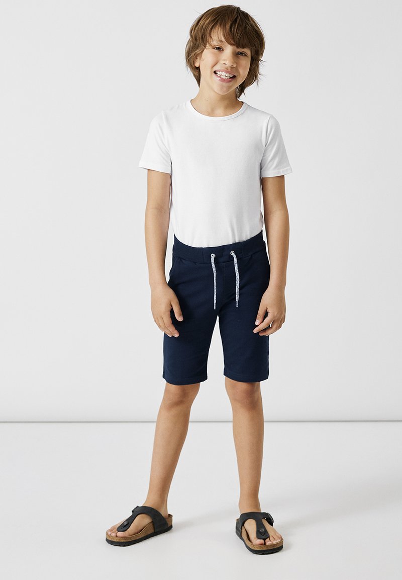 Vit kortärmad T-shirt, marinblå shorts med dragsko, och svarta flip-flops. Enkel design, lättviktig tyg, avslappnad stil.