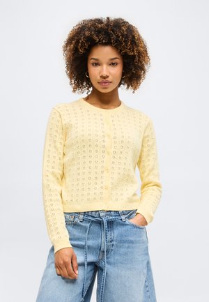 Kobieta o kręconych włosach, ubrana w bladożółty sweter na guziki i jasnoniebieskie, wysokie jeansy, z jedną ręką w kieszeni na białym tle.