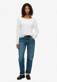 Witte lange mouwen shirt, losse blauwe jeans, zwarte riem en donkere instappers met witte sokken; model staat tegen een effen lichte achtergrond.