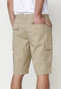 Shorts cargo bej cu față plată, buzunare laterale și stofă texturată. Prezintă un loop pentru curea și un buzunar pe spate.