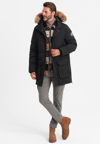 Schwarze Daunenjacke mit einer mit Fell gefütterten Kapuze, mit Reißverschluss vorne, großen Taschen und einem lässigen Schnitt, getragen über einem gemusterten Hemd und einer Hose.