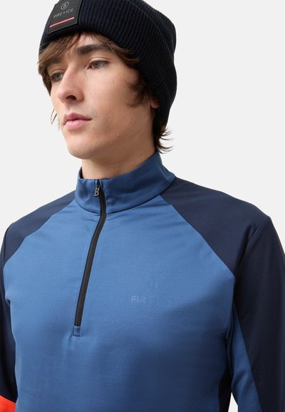 Blue athletic half-zip pullover with navy raglan sleeves, smooth texture, logo on chest, complemented by a black ribbed beanie.