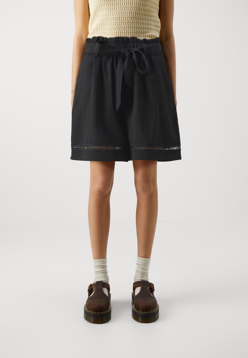 Femme portant un short noir taille haute avec une ceinture nouée, un haut en tricot sans manches beige, des chaussettes blanches et des chaussures à plateforme marron avec boucle.