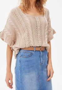 Suéter recortado de punto color beige con patrones de trenzado, mangas amplias, combinado con una falda de mezclilla de cintura alta y un cinturón marrón.