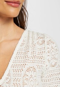 Décolleté et épaule d'une femme dans un vêtement en crochet blanc avec des motifs complexes, lèvres brillantes et longs cheveux foncés visibles.