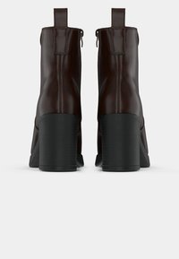 Um par de botas de cano curto em couro castanho escuro com saltos altos pretos, mostradas de trás contra um fundo branco liso.