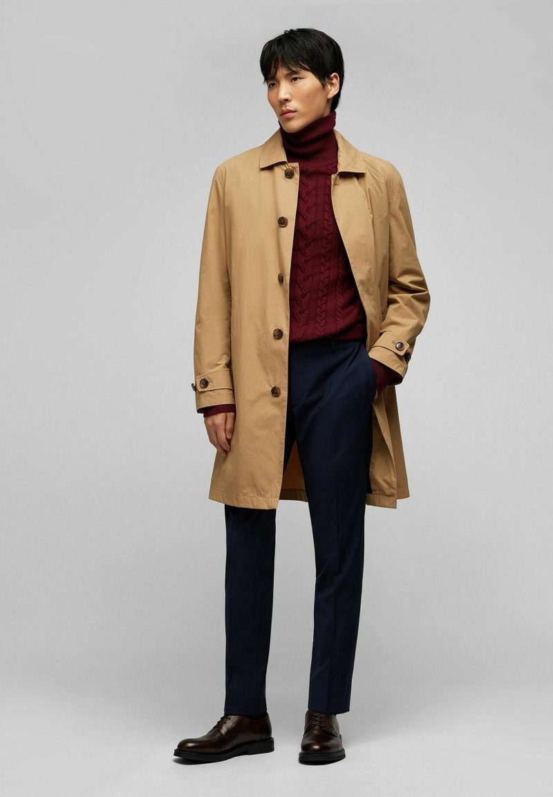 Manteau beige avec boutons marron sur un col roulé en tricot bordeaux, associé à un pantalon marine et des chaussures en cuir marron foncé à lacets.