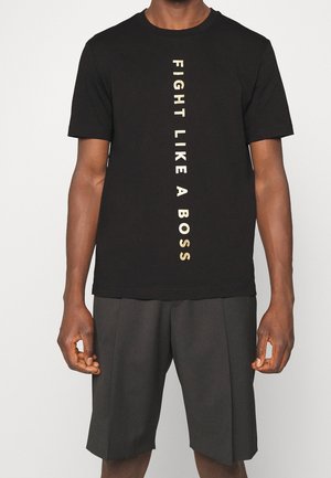 T-shirt en coton noir avec un texte doré vertical qui dit "BATTEZ-VOUS COMME UN PATRON." Manches courtes et coupe décontractée. Associé à un short sombre.