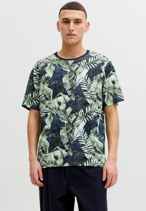 Joven con camiseta azul marino de manga corta con patrones de hojas tropicales verdes y azules, de pie contra un fondo claro y liso.