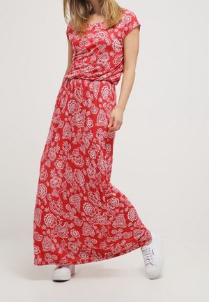 Žena v červených květovaných maxi šatech s krátkým rukávem a bílých teniskách stojí před jednoduchým pozadím.