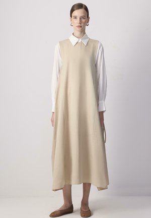 Beige ærmeløs kjole over en hvid skjorte med krave, flydende A-line form, blødt stof og minimalistisk design, parret med brune flade sko.