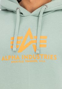 Zöld kapucnis pulóver puha anyagból, narancssárga grafikai szöveggel "ALPHA INDUSTRIES" és logóval, zsinórokkal és fém megállítókkal.