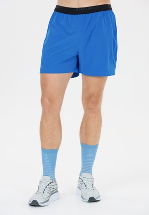 Mann trägt blaue Sportshorts, hellblaue Kniestrümpfe und weiße Laufschuhe, steht vor weißem Hintergrund.