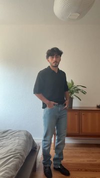 Črna kratka srajca s kratkimi rokavi, svetloplavi jeans in črne čevlje. Samostojna figura s rastlino in lesenim pohištvom v ozadju.