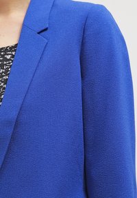 Světle modrý blazer s texturovanou tkaninou, zapínáním na jeden knoflík a přiléhavým střihem s ostrými klopami a čistou siluetou.