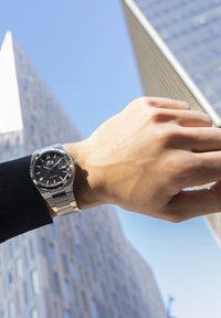 Reloj de acero inoxidable con un dial negro texturizado, marcadores plateados y una pulsera de eslabones, exhibido sobre una mano con un fondo de cielo azul.