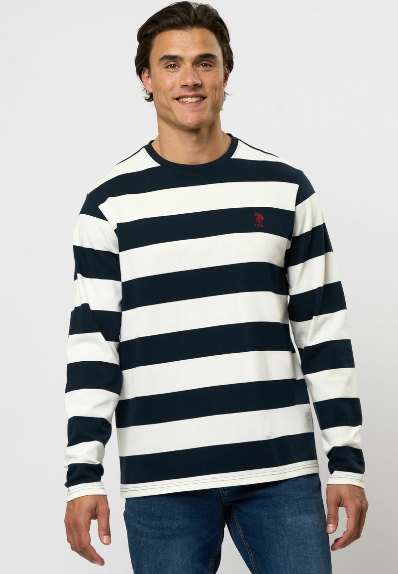 Man draagt een zwart-wit gestreept shirt met lange mouwen en een rood logo, blauwe spijkerbroek, glimlacht en staat voor een effen lichte achtergrond.