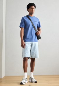 Blauwe katoenen t-shirt met ronde hals, lichte spijkerbroekshorts, en witte sokken in combinatie met grijze New Balance sneakers. Tasband over de schouder.
