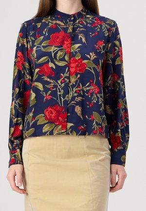 Blouse bleu marine à imprimé floral rouge et manches longues associée à une jupe beige, portée par une personne vue du cou aux hanches.