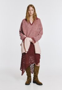 Prevelik ročno pleten pulover roza barve z bordo poudarki, nošen prek bordo čipkaste obleke, kombiniran z rjavimi škornji do kolen.