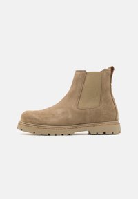 Botină Chelsea din suede maro, cu panouri elastice pe laterale, vârf rotund și talpă din cauciuc texturat, având modele de traction pentru aderență.