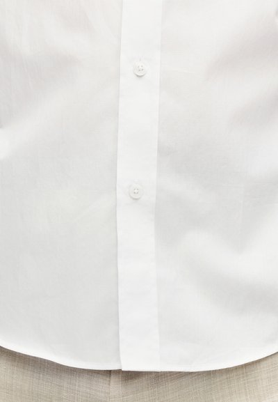 Λευκό πουκάμισο με κουμπιά, μπαίνει μέσα σε μπεζ παντελόνι από υφή υφάσματος, εστίαση στη μεσαία περιοχή και στις λεπτομέρειες των κουμπιών.