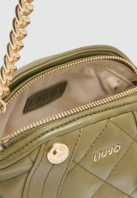 Olijf groene gequilt leren tas met een gouden schouderband, ritssluiting en een gouden logo-accent op een beige voering.