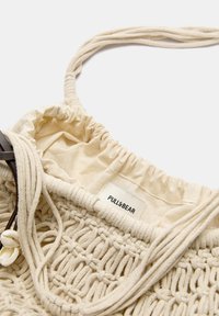 Borsa intrecciata beige con chiusura a coulisse, materiale in cotone e etichetta del marchio. Le cinghie sono realizzate in tessuto attorcigliato.