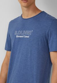 Niebieska koszulka z krótkim rękawem, uszyta z miękkiej tkaniny, z okrągłym dekoltem. Posiada biały nadrukowany tekst: "S. OLIVER Elevated Casual."