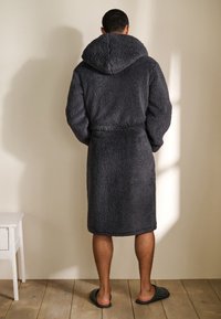 Donkergrijze pluche badjas met capuchon, gestructureerde stof, gestrikt in de taille, kniehoogte, gedragen met zwarte gestreepte slippers op een houten vloer.