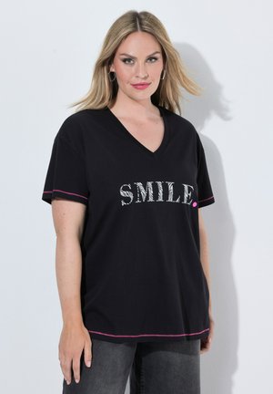 Blonde vrouw die een zwart V-hals T-shirt draagt met de tekst "SMILE." en roze stiksels, staand tegen een lichtgrijze achtergrond.