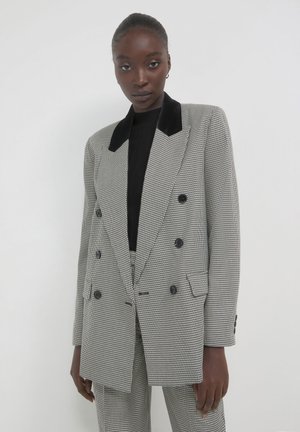 Donna che indossa un blazer doppiopetto in bianco e nero a scacchi, con colletto in velluto nero, pantaloni abbinati e una maglietta nera, in piedi contro una parete bianca.