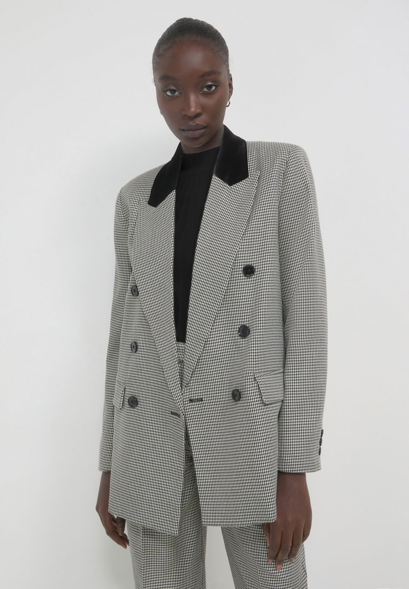 Donna che indossa un blazer doppiopetto in bianco e nero a scacchi, con colletto in velluto nero, pantaloni abbinati e una maglietta nera, in piedi contro una parete bianca.