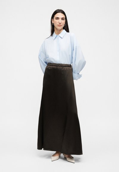 Γυναίκα που στέκεται, φορώντας ένα ανοιχτό μπλε φαρδύ πουκάμισο με κουμπιά, σκούρα καφέ μακριά φούστα και κρεμ μυτερά τακούνια σε απλό φόντο.