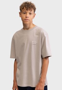 Beige cotton t-shirt with short sleeves, crew neck, and a small graphic logo on the left chest area. Relaxed fit, smooth texture.