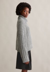 Szary sweter z dzianiny z wysokim kołnierzem, żebrowaną strukturą i opadającymi ramionami, w połączeniu z ciemną spódnicą. Gładkie tło.