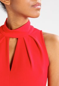 Haut rouge sans manches avec un col haut orné de détails plissés et une ouverture en forme de goutte, confectionné dans un tissu lisse à la texture légèrement brillante.