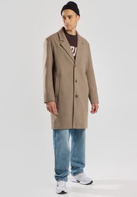 Cappotto in misto lana marrone con colletto a punta, due tasche frontali e chiusura a tre bottoni, indossato sopra una maglietta scura e jeans.