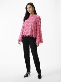 Pink fringe sweater with wave pattern, loose fit, long sleeves, paired with black tailored pants and black heeled mules. Simple background.