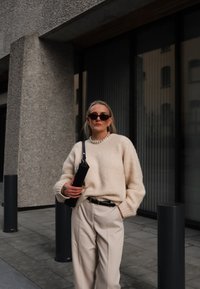 Beige, texturerad tröja med vida ärmar, i kombination med ljusbeiga byxor. Tillbehör inkluderar en svart axelväska och mörka solglasögon.