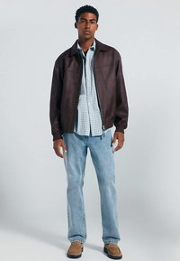 Giacca di pelle marrone con zip, indossata sopra una camicia a quadri azzurro chiaro, abbinata a jeans azzurri e scarpe marroni.