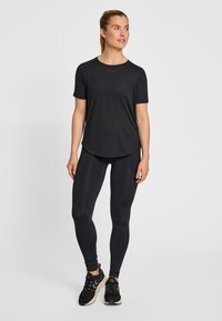 Kobieta stojąca, ubrana w czarną koszulkę z krótkim rękawem, czarne legginsy i czarne trampki z białymi podeszwami, patrząca lekko w prawo.