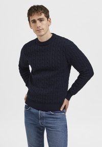 Marineblauer Strickpullover mit strukturiertem Zopfmuster, Rundhalsausschnitt und gerippten Bündchen und Saum. Kombiniert mit hellblauen Jeans.